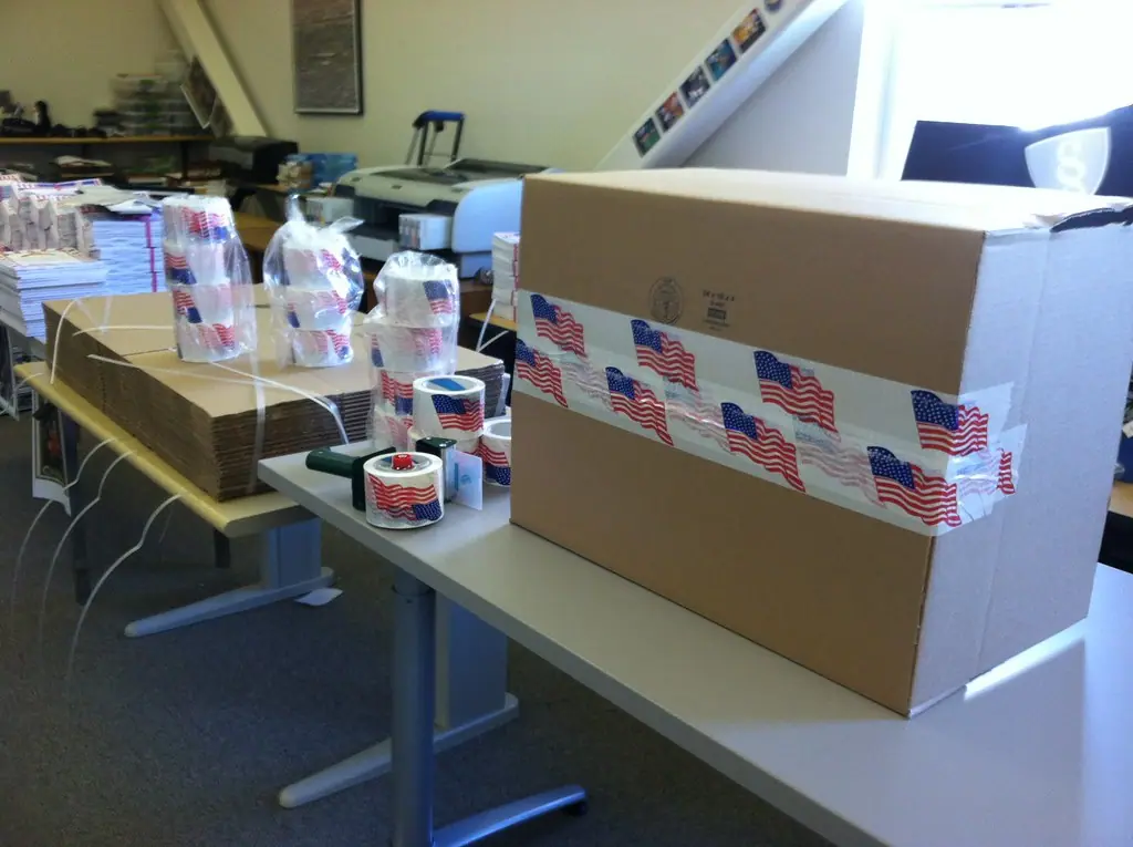 A carton sealed with printed packing tape, rolls of tape on the table behind